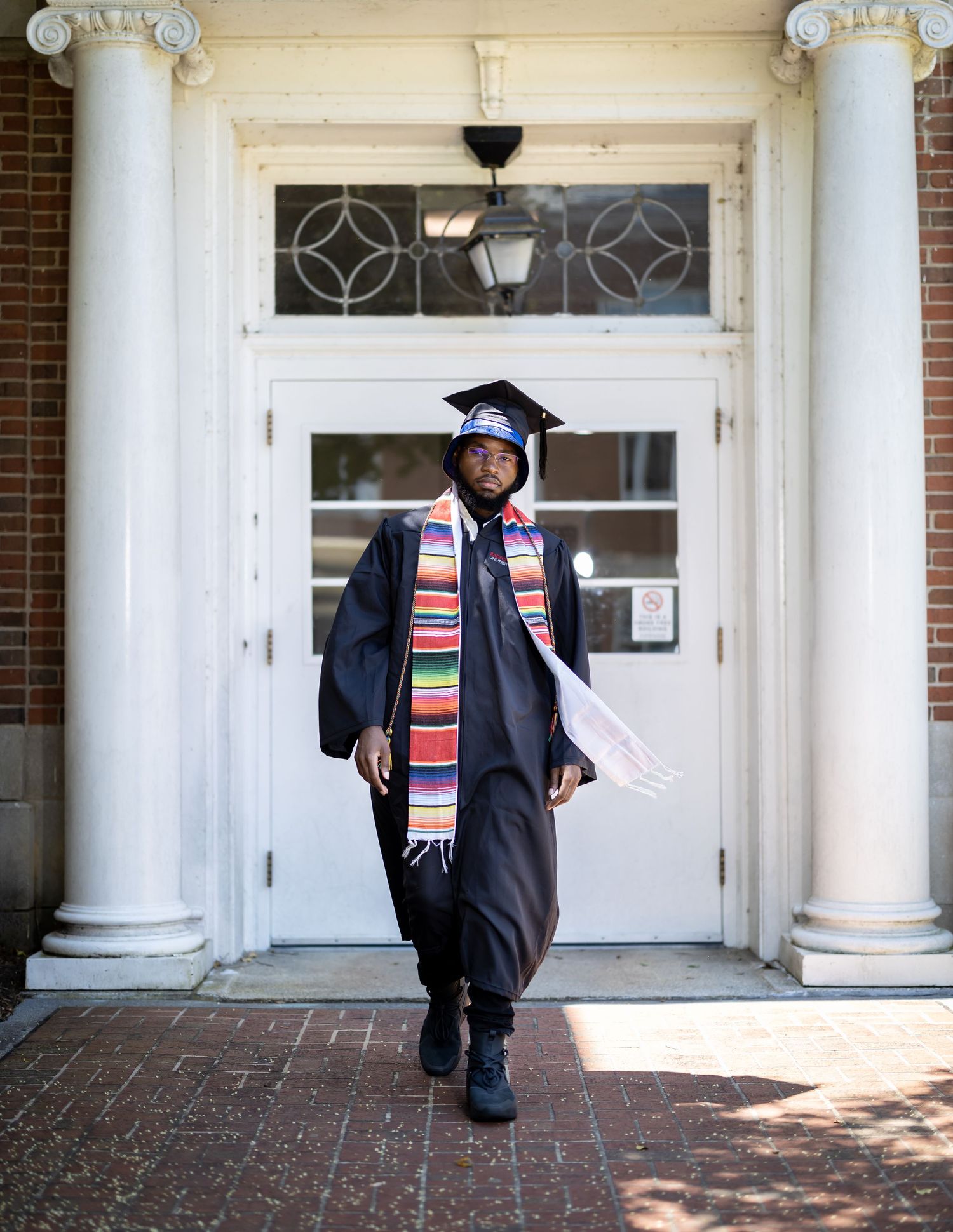 Graduate in academic regalia with colorful stole walking through columned entrance of building.