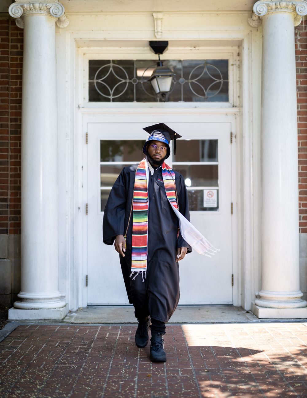Graduate in academic regalia with colorful stole walking through columned entrance of building.
