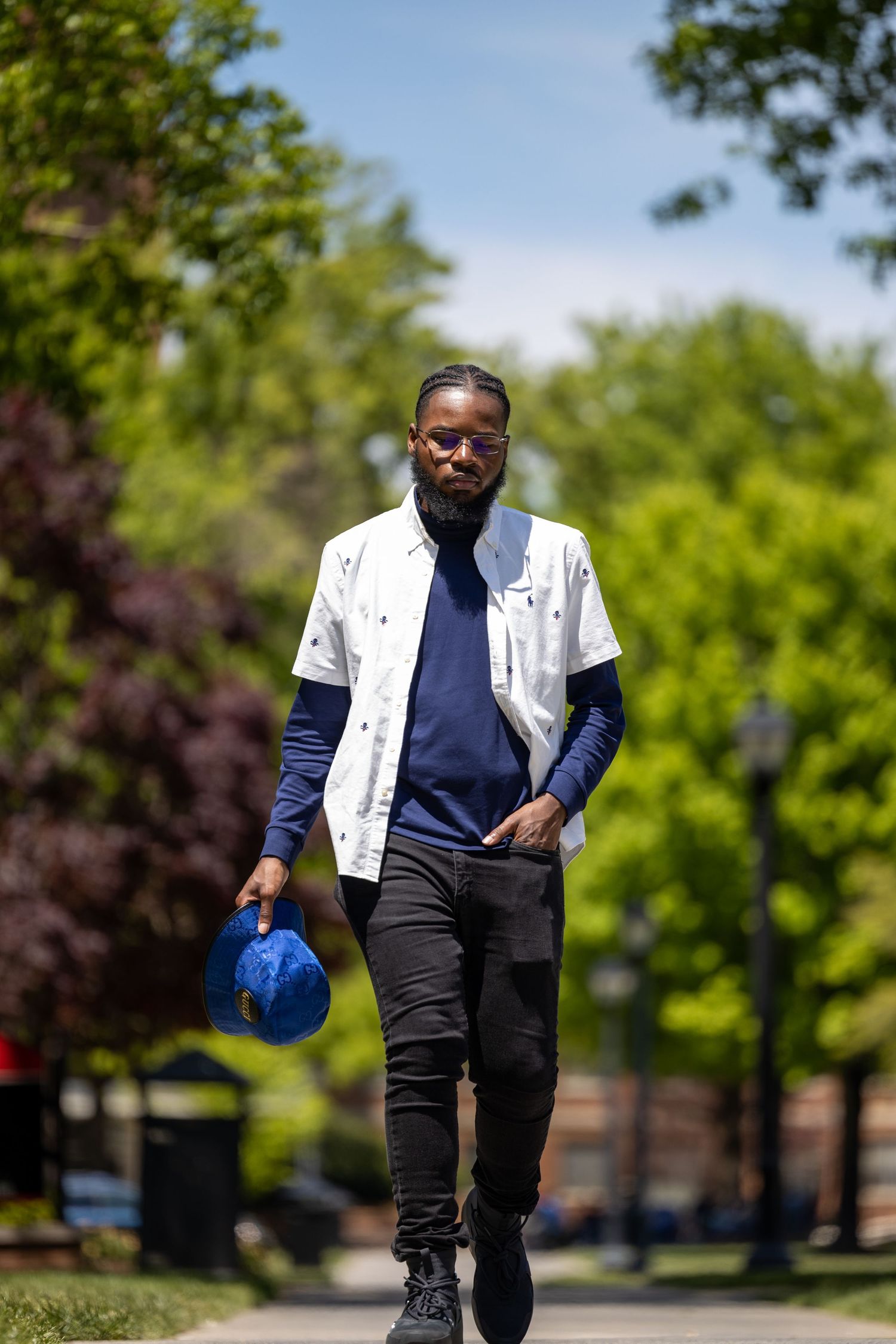 Person in white jacket walking along tree-lined path holding blue cap on sunny day.