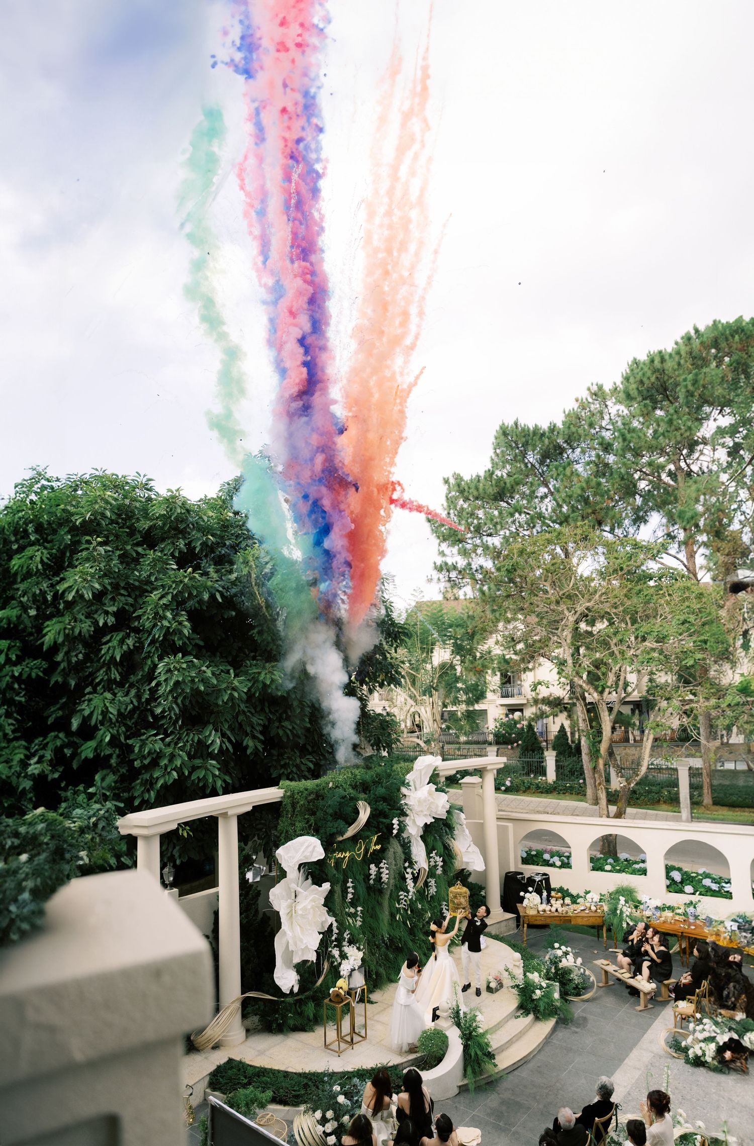 Colorful smoke trails burst into the sky above a white-decorated outdoor venue with trees and parked cars visible below.
