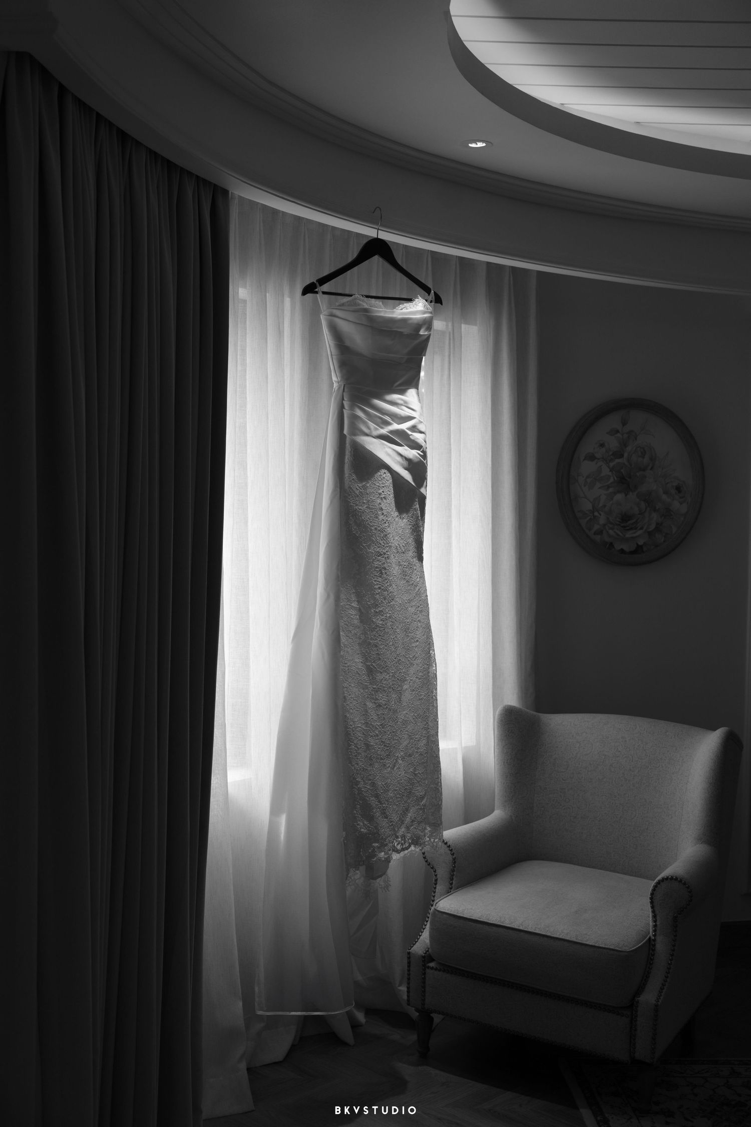 An elegant white wedding dress hangs illuminated in front of a window next to an armchair in a dramatic black and white photo.