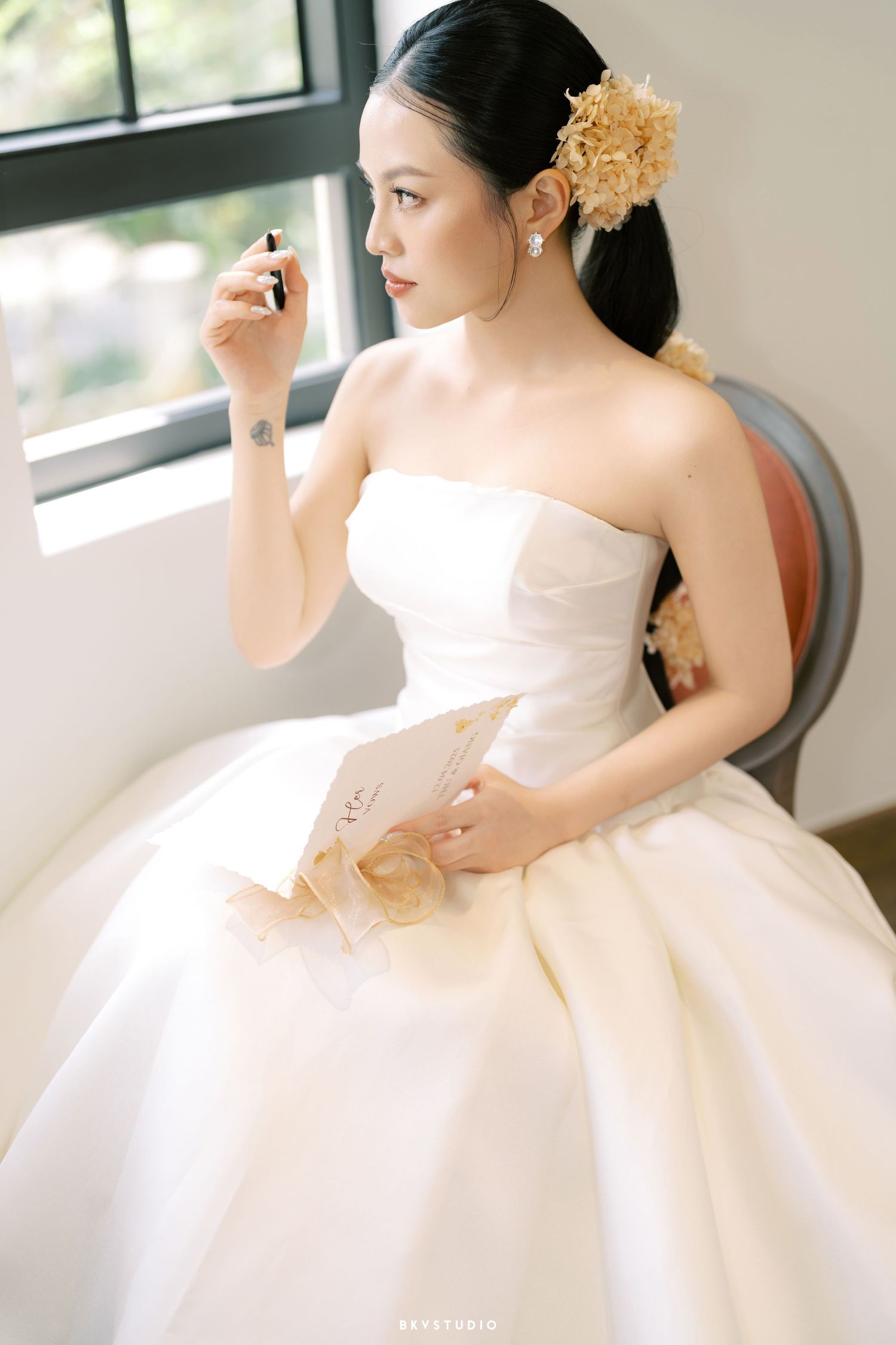 A bride in an elegant strapless white ball gown gazes out a window while holding a note.