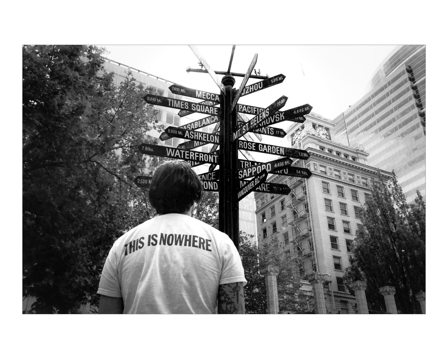 Black and white photo showing a person looking up at a multidirectional street sign post in an urban setting.