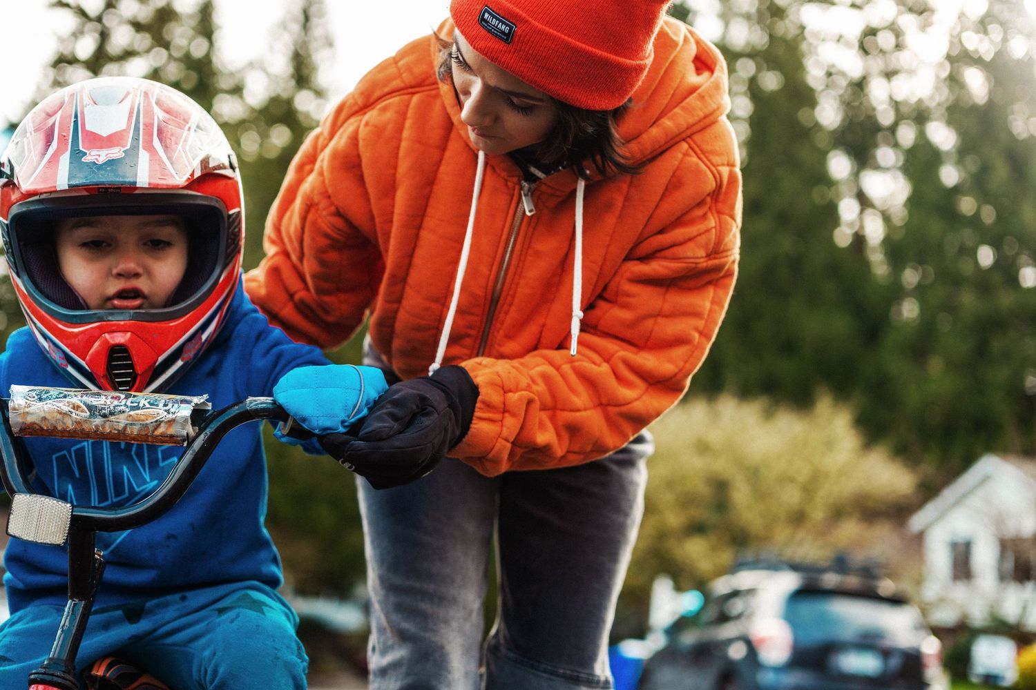 Person in orange hoodie helps child wearing blue motorcycle helmet and protective gear with outdoor activity.