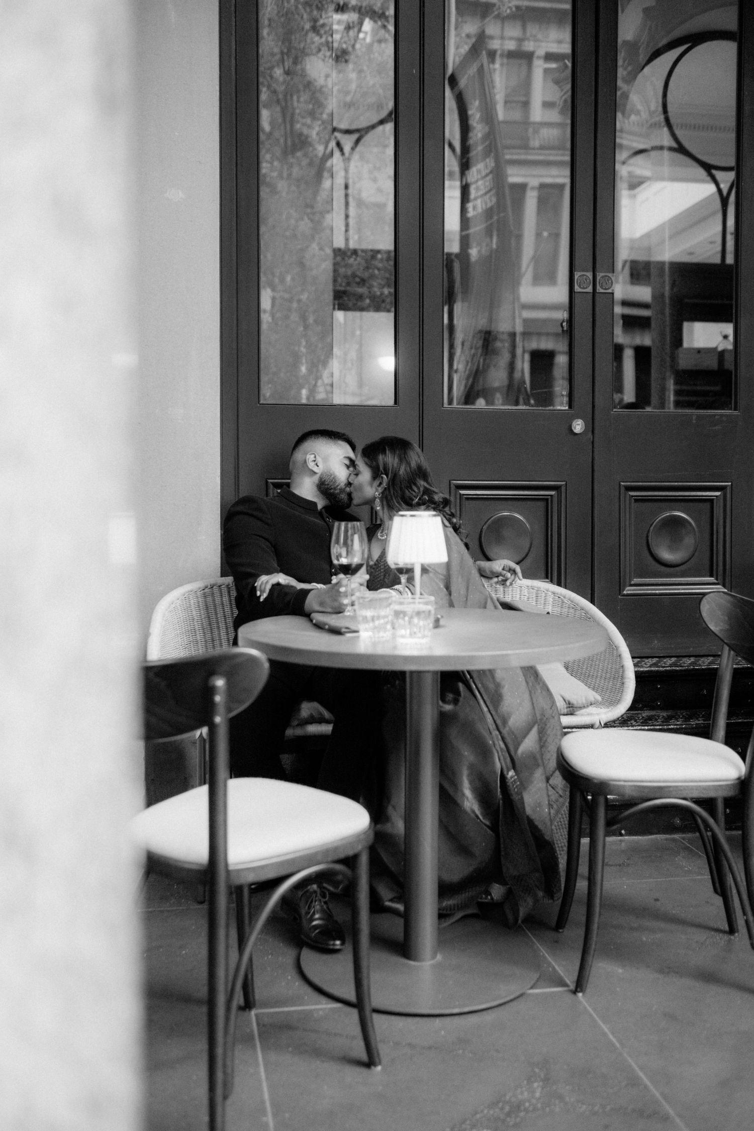 A romantic black and white photograph of a couple sharing an intimate moment at a small cafe table outdoors.