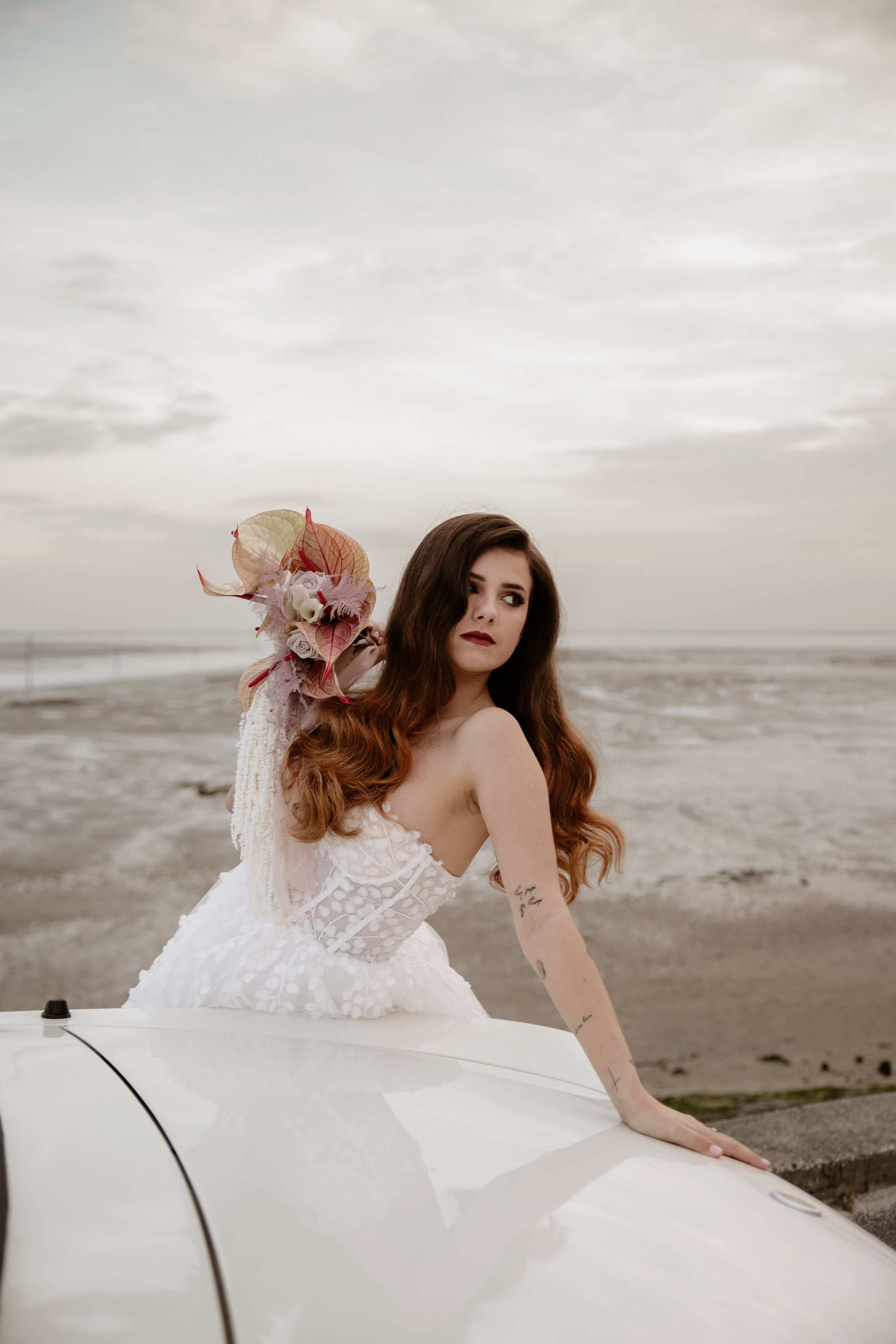 Bride with floral hairpiece poses on vintage white car by oceanside.
