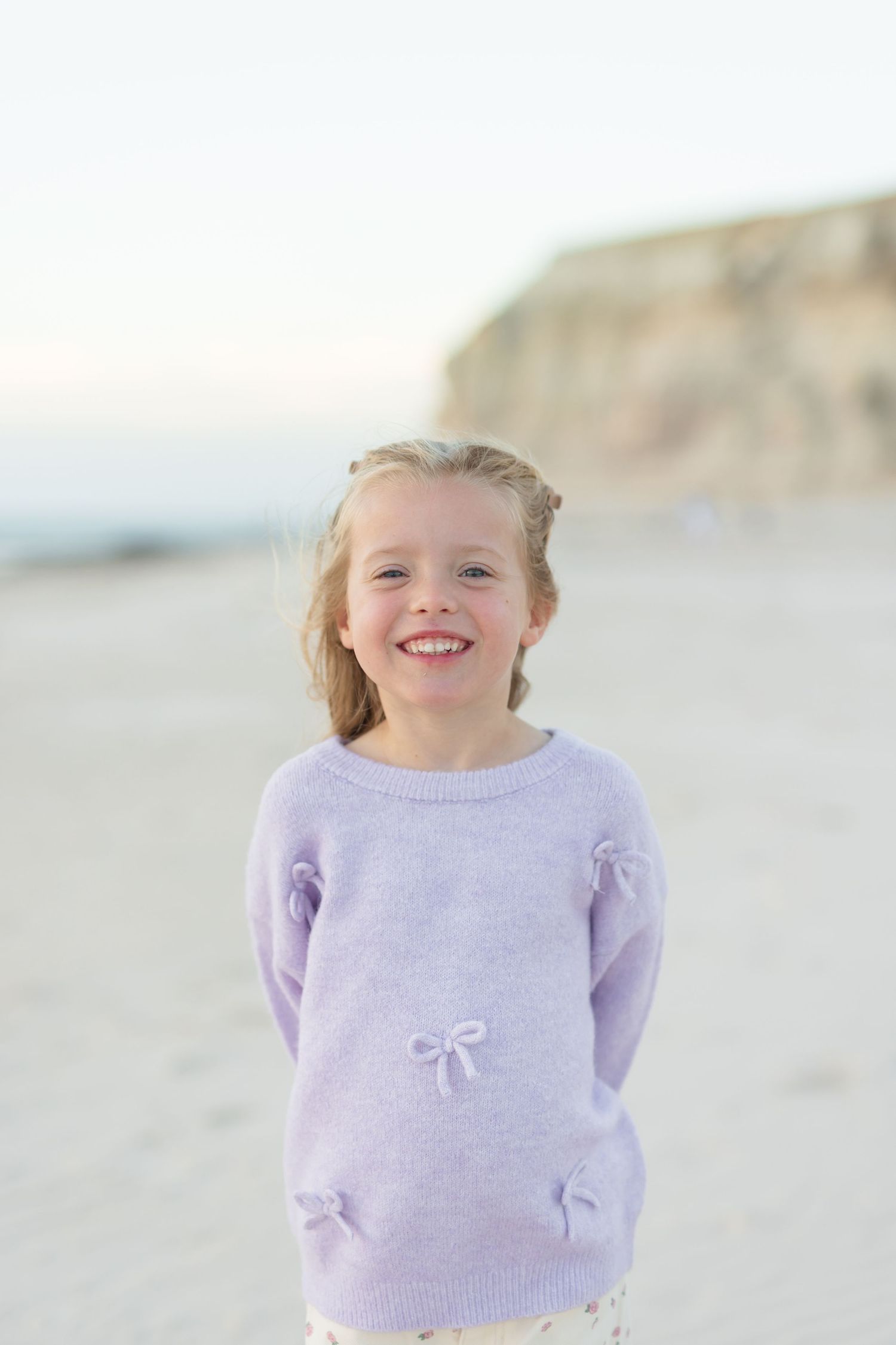 Smiling young girl in a lavender bow sweater standing on a sandy beach with cliffs in the background.