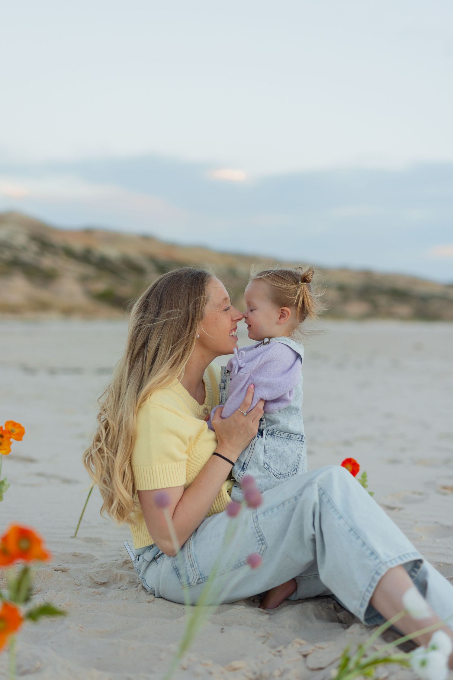 A mother and toddler share a tender nose-to-nose moment while sitting on a sandy beach surrounded by colorful flowers.