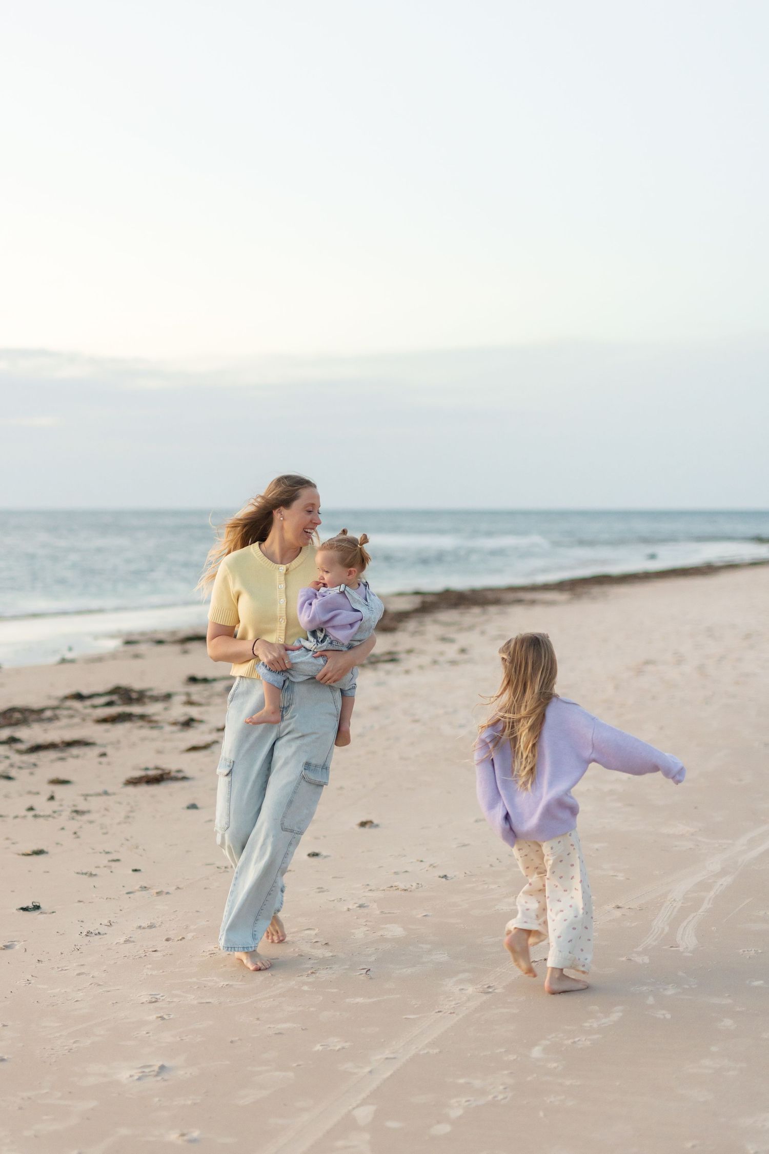 A mother walks barefoot on a sandy beach at sunset, carrying a baby while her young daughter runs alongside her.
