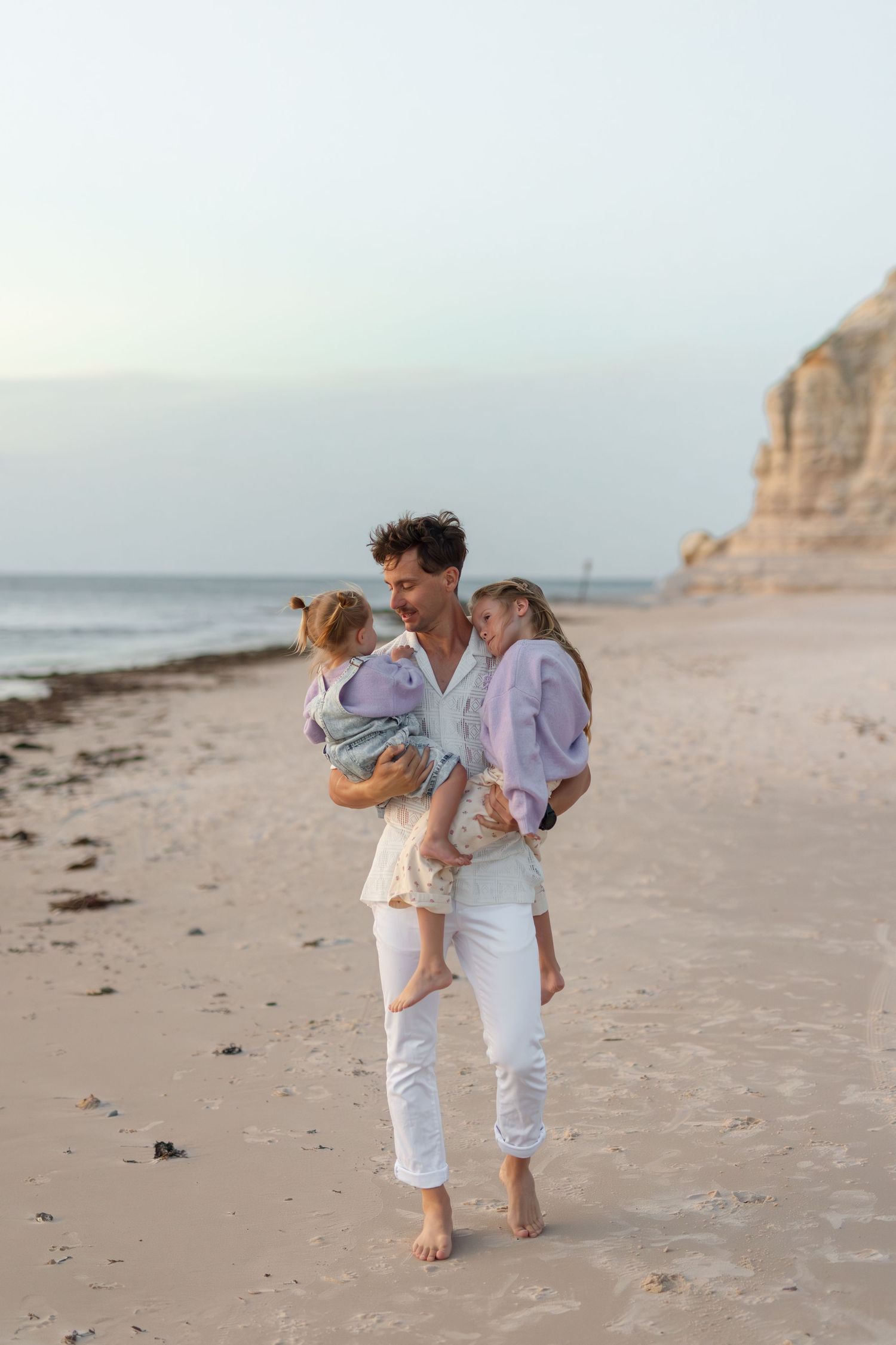 A man carries two young children on a sandy beach at sunset, with cliffs and calm ocean in the background.