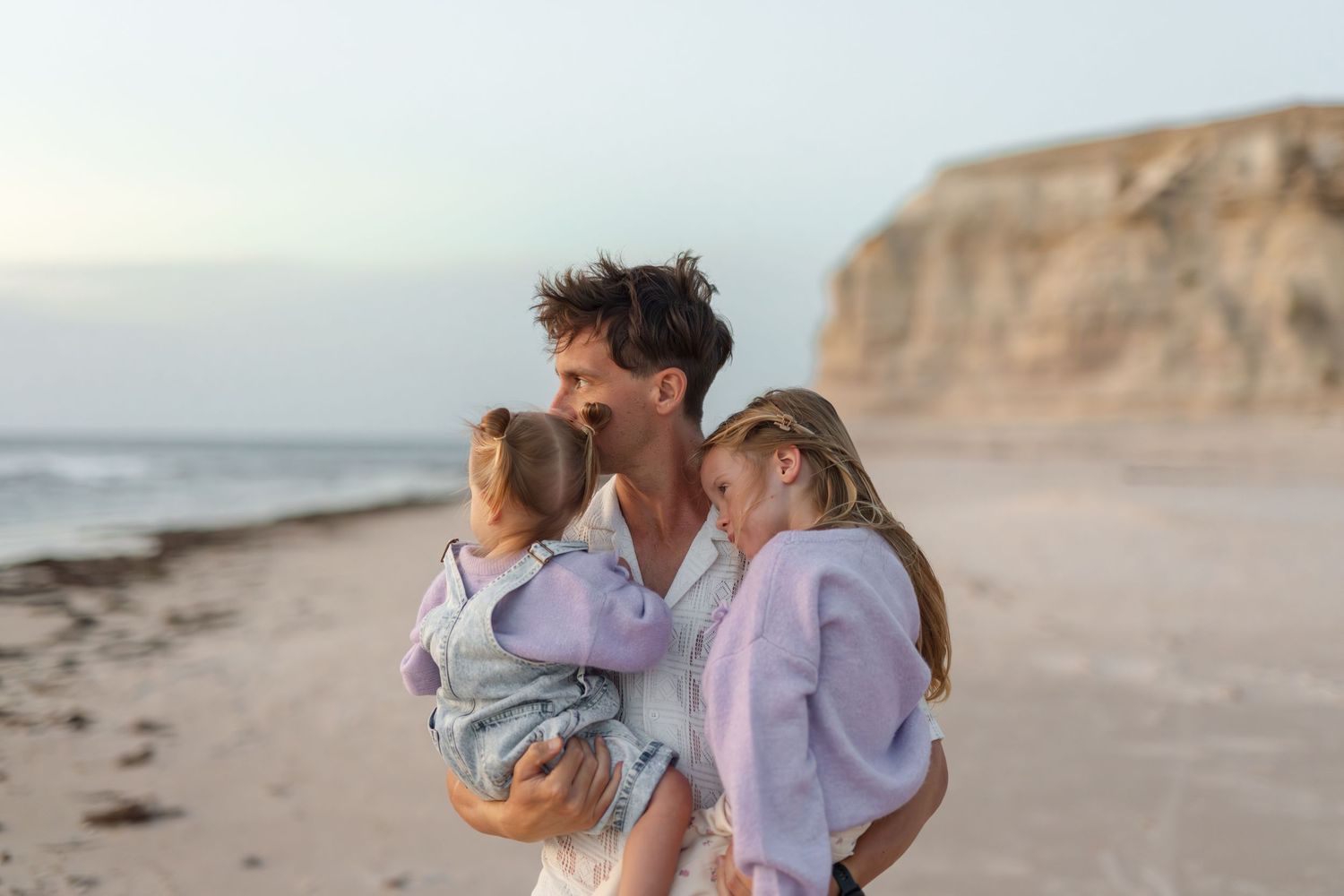 A father holds his two young daughters on a sandy beach at dusk with ocean waves and cliffs behind them.