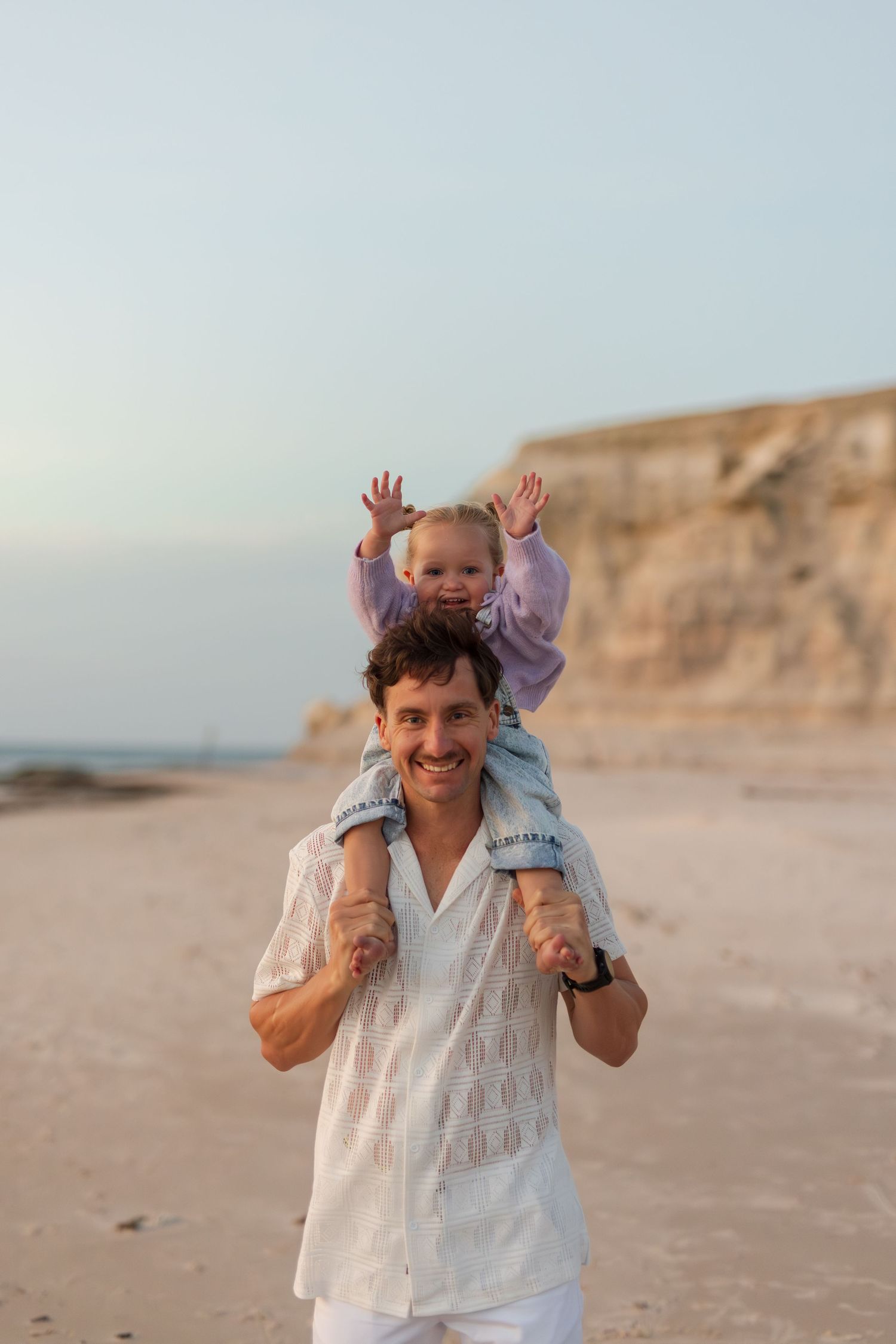 A smiling father carries his happy toddler on his shoulders while walking along a scenic sandy beach at sunset.