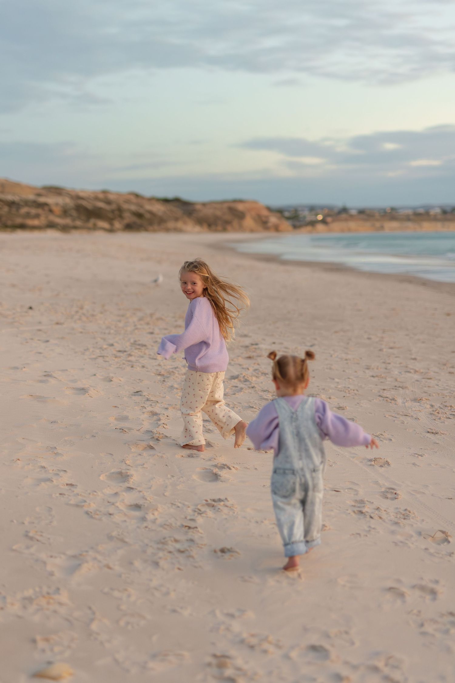 Two young girls in purple sweaters run barefoot on a sandy beach at sunset with ocean waves in the background.