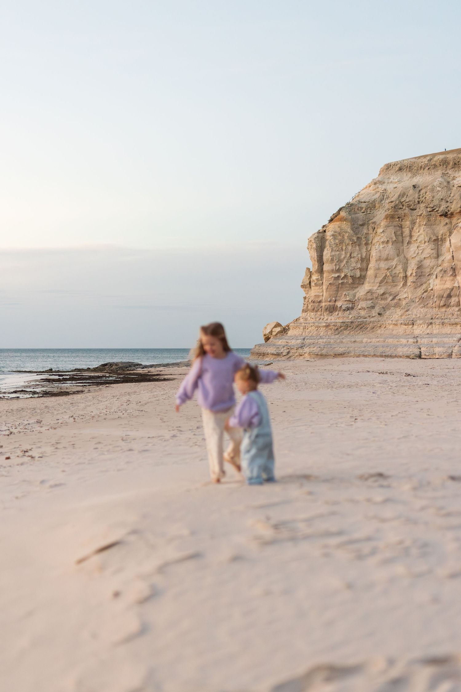 Two little girls in matching purple sweaters walk along a sandy beach near dramatic coastal cliffs at dusk.