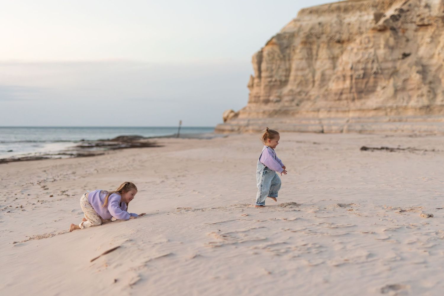 Two young children playing on a sandy beach at sunset with rocky cliffs in the background.