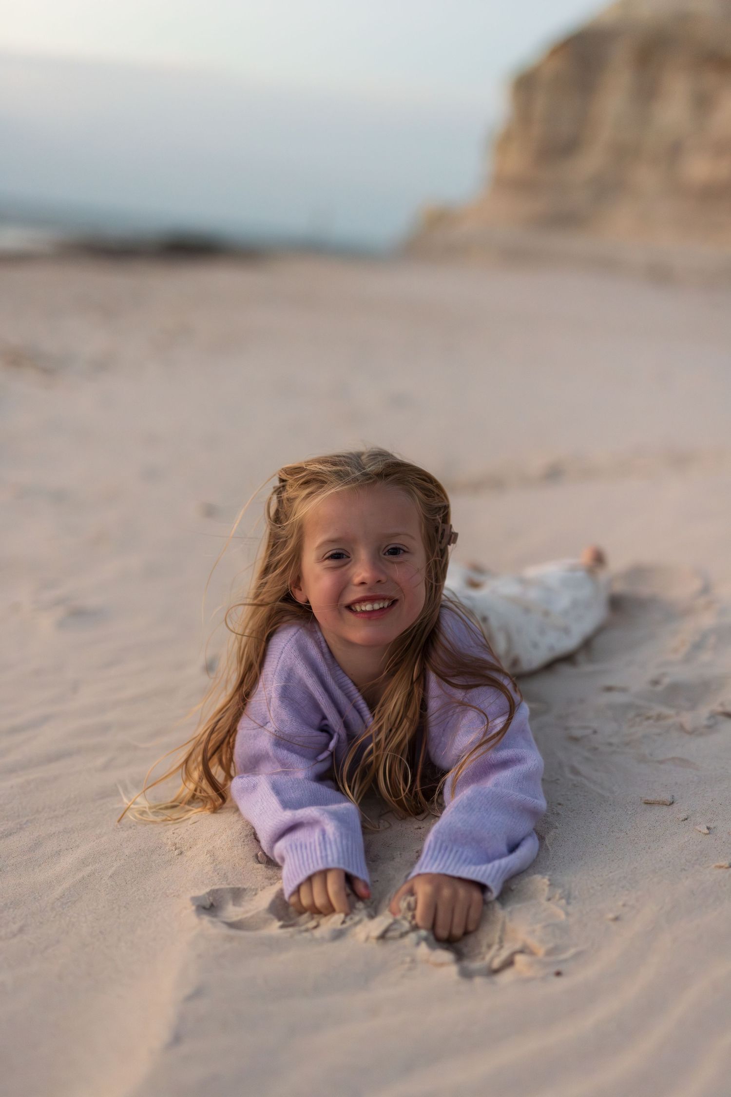 Young girl with long blonde hair smiling while lying on sandy beach wearing a purple sweater.