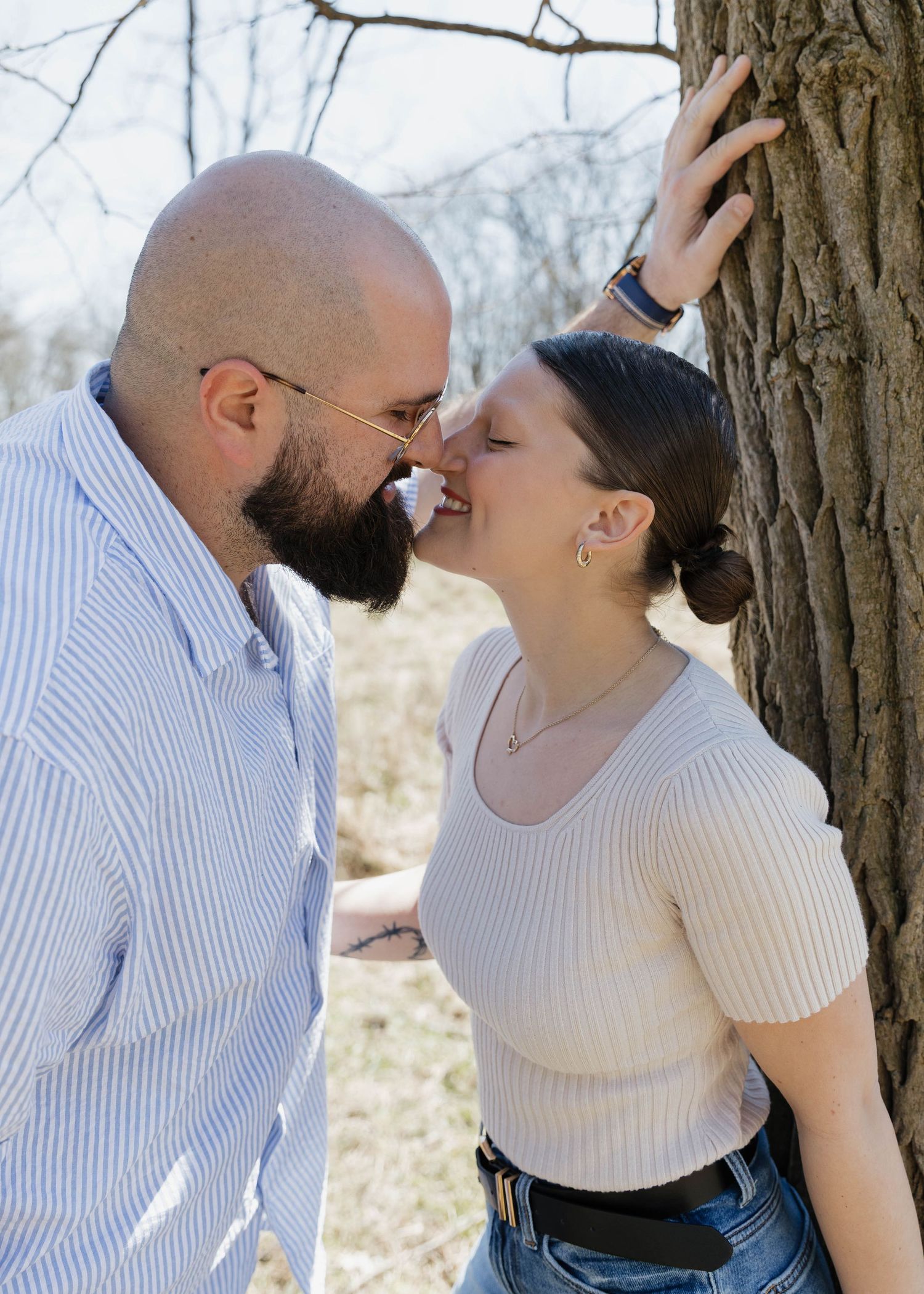 A romantic couple share a tender moment together against a tree trunk in an outdoor setting.