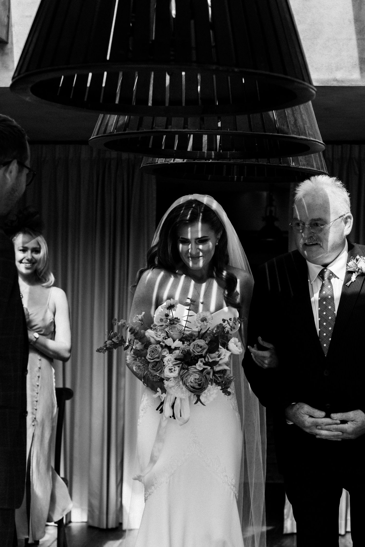 Black and white photo of a bride walking down the aisle with her father, holding a floral bouquet at a wedding ceremony.