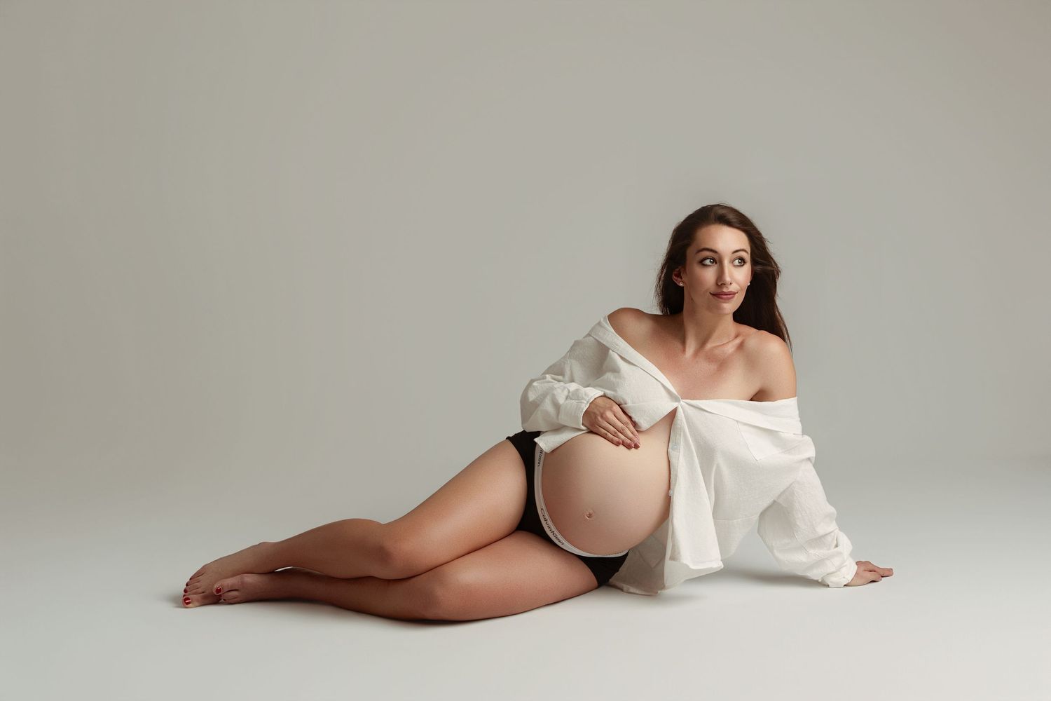 Pregnant woman in black underwear and open white shirt sitting on floor, cradling baby bump in elegant maternity photo.