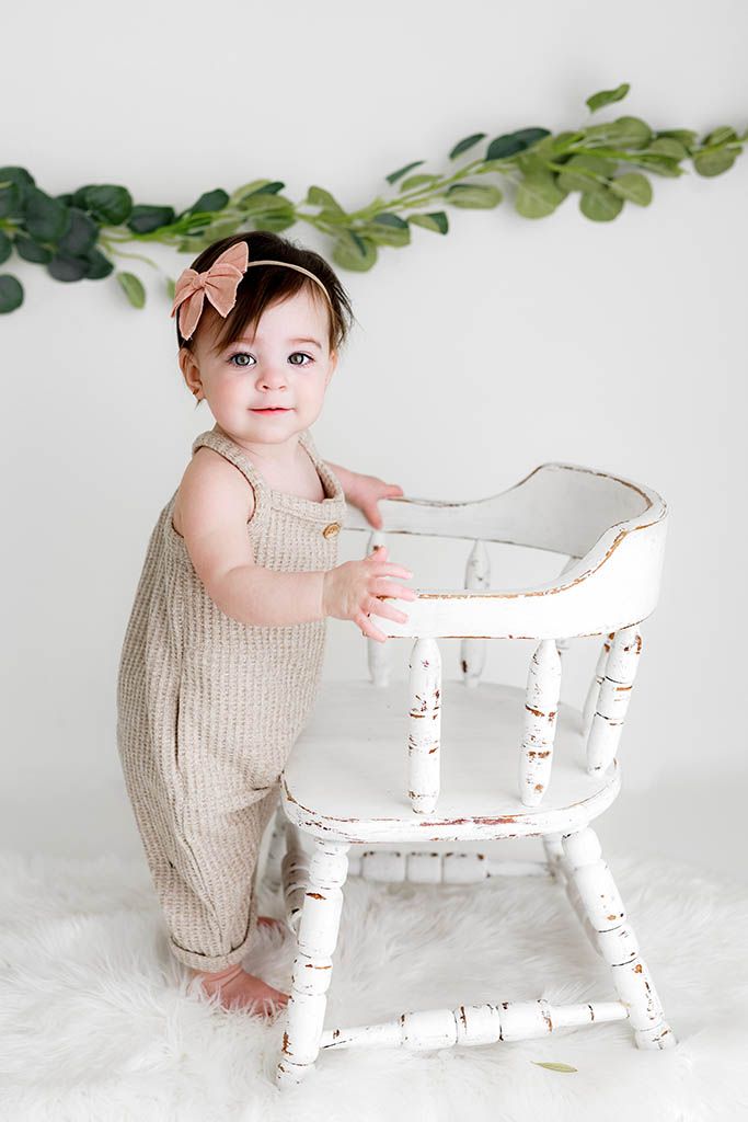 Adorable baby girl in beige romper and bow headband standing beside a vintage white wooden high chair in a studio.