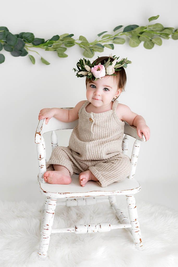 Adorable baby in knit romper and floral crown sits in white rustic chair with greenery garland backdrop.