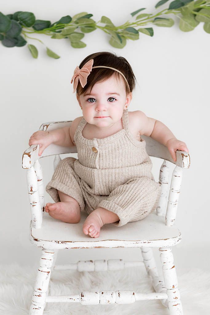 Adorable baby girl in beige knit romper and bow headband sits on a rustic white chair with greenery backdrop.