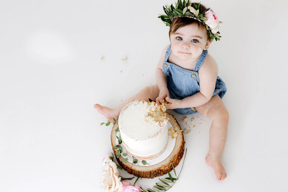 Baby girl in denim romper and floral crown smashes white two-tier cake on white background.
