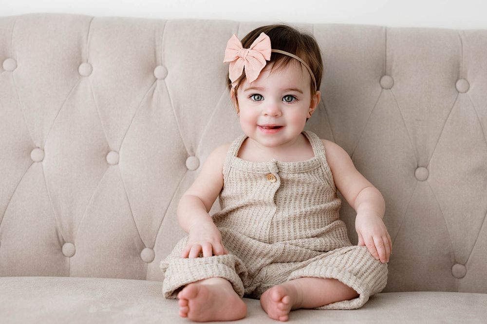 Smiling baby girl in knit romper and pink bow headband sitting on a beige tufted sofa.
