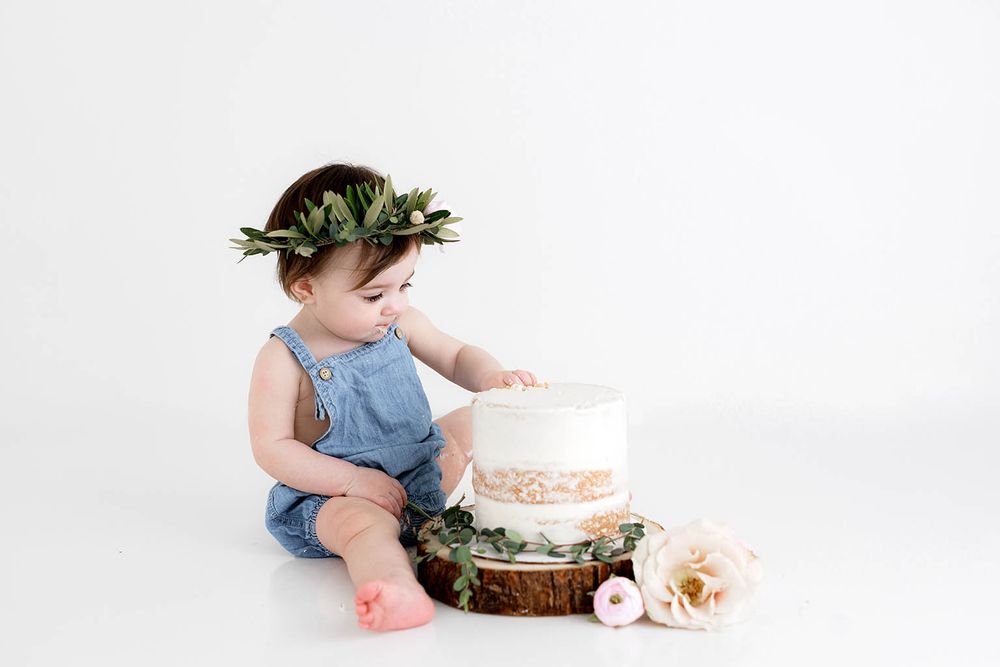 Baby girl in denim romper and floral crown reaching into a white first birthday smash cake.