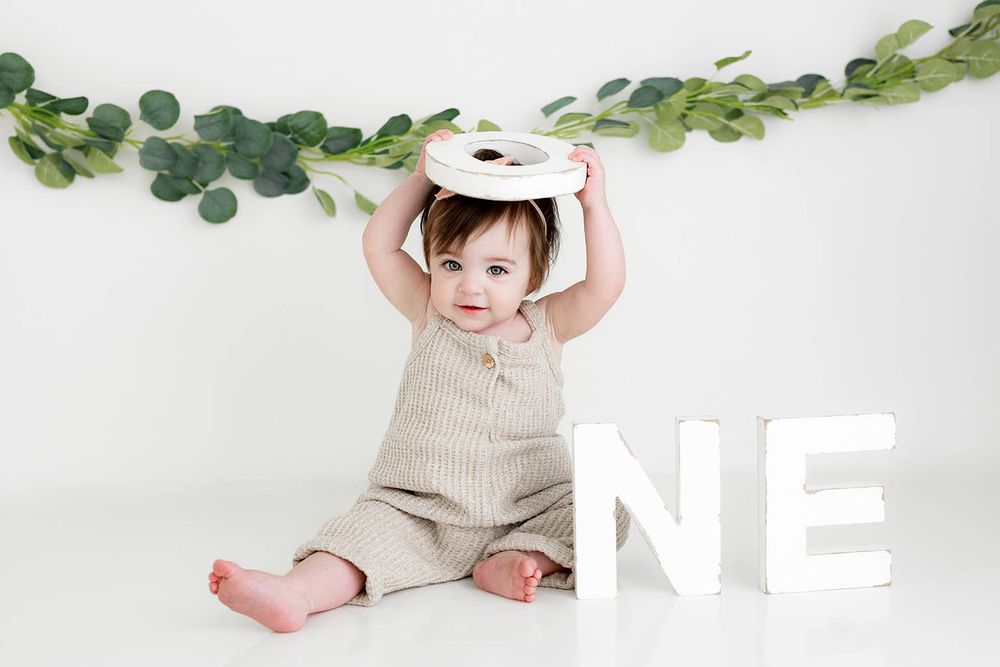A smiling baby in a beige romper holds a round object overhead beside ONE letters with eucalyptus garland backdrop.