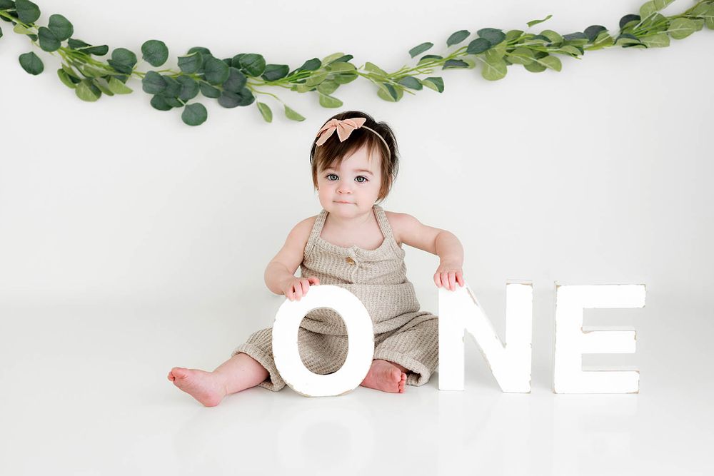 Adorable baby girl in tan romper sitting with white ONE letters and eucalyptus garland backdrop for first birthday photo.