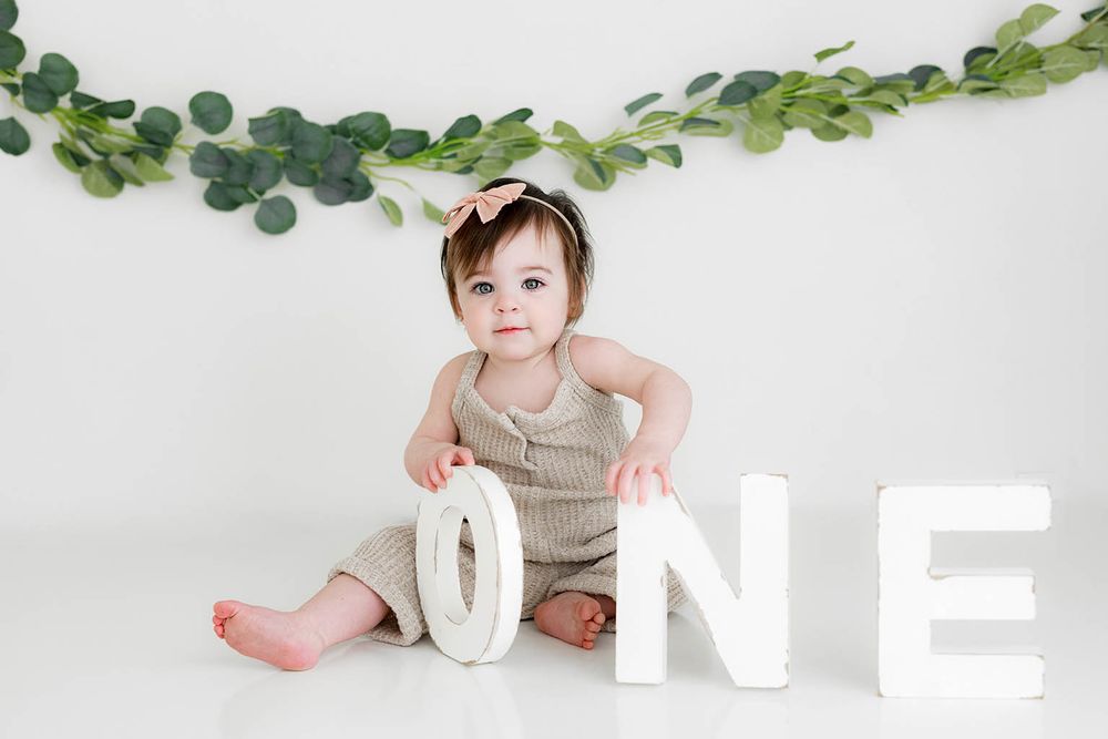 Baby girl in romper sits with ONE letters for first birthday cake smash photo session.
