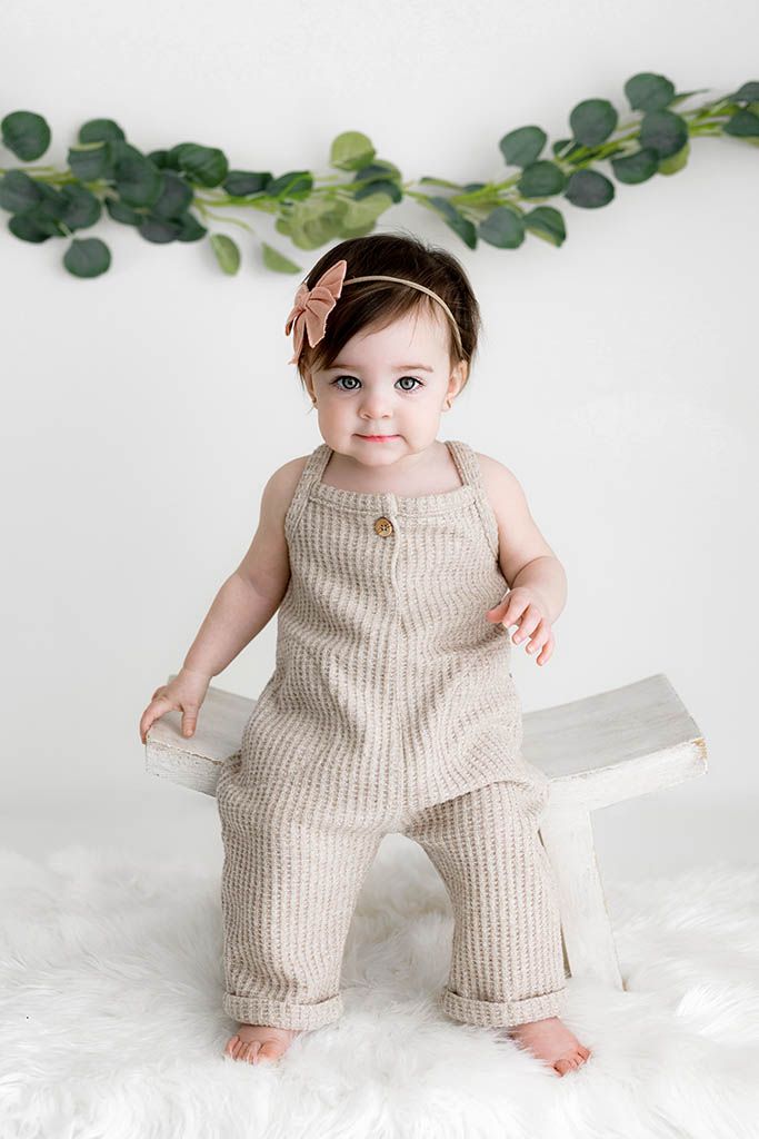 Adorable baby girl in striped linen overalls and bow headband sitting on white bench with eucalyptus backdrop.