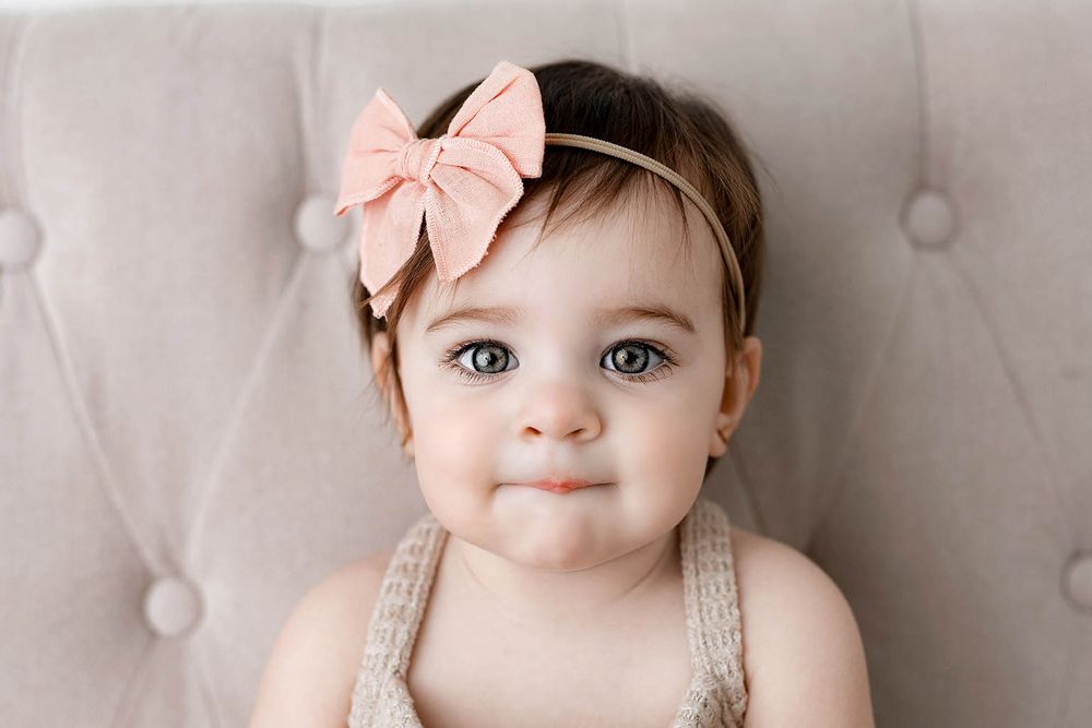 Adorable baby girl with big blue eyes wearing a pink bow headband and beige romper, posed on a tufted gray background.