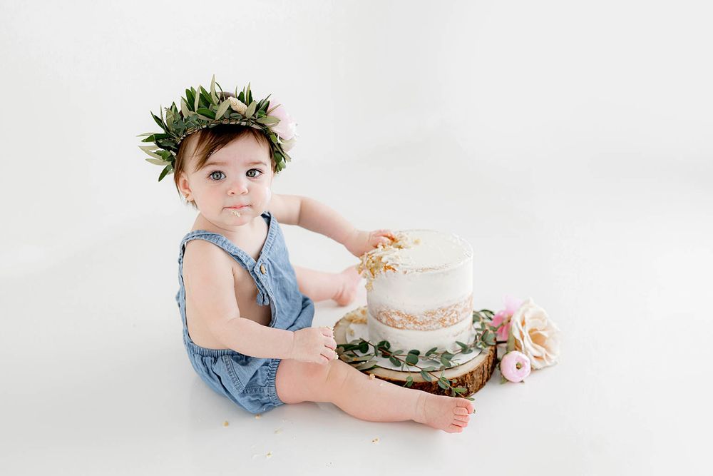 Cute baby in blue romper wearing flower crown sitting beside a white birthday smash cake.