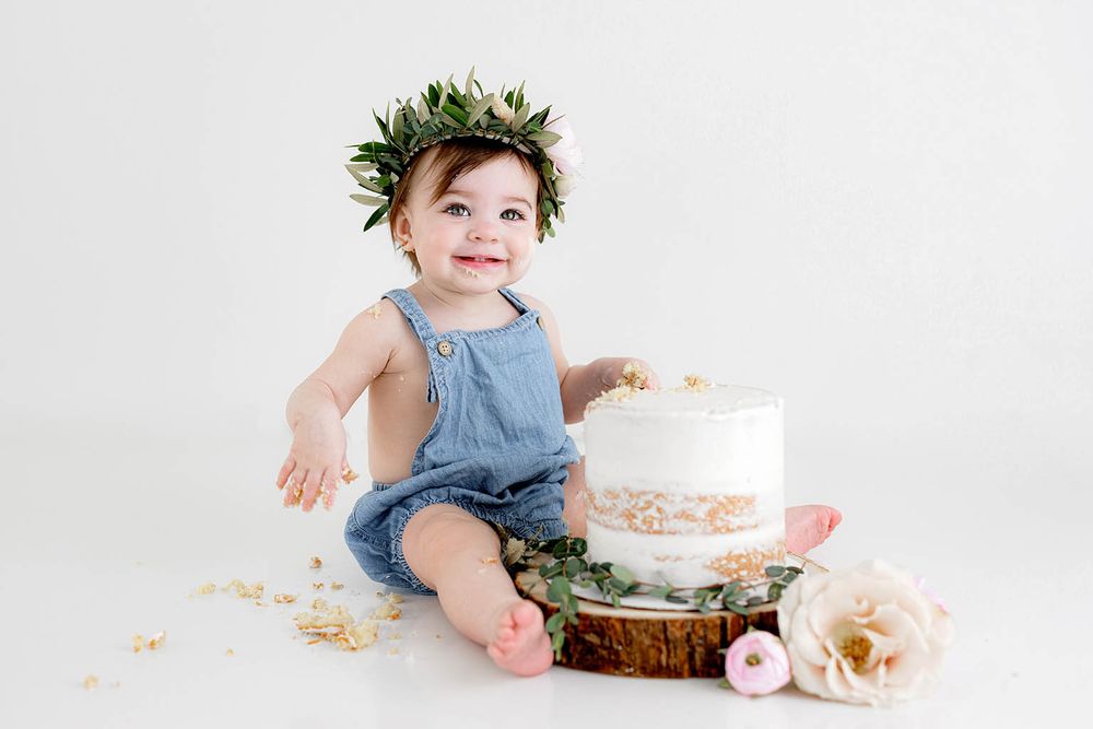 Happy baby girl in denim overalls and floral crown sitting next to a white smash cake.