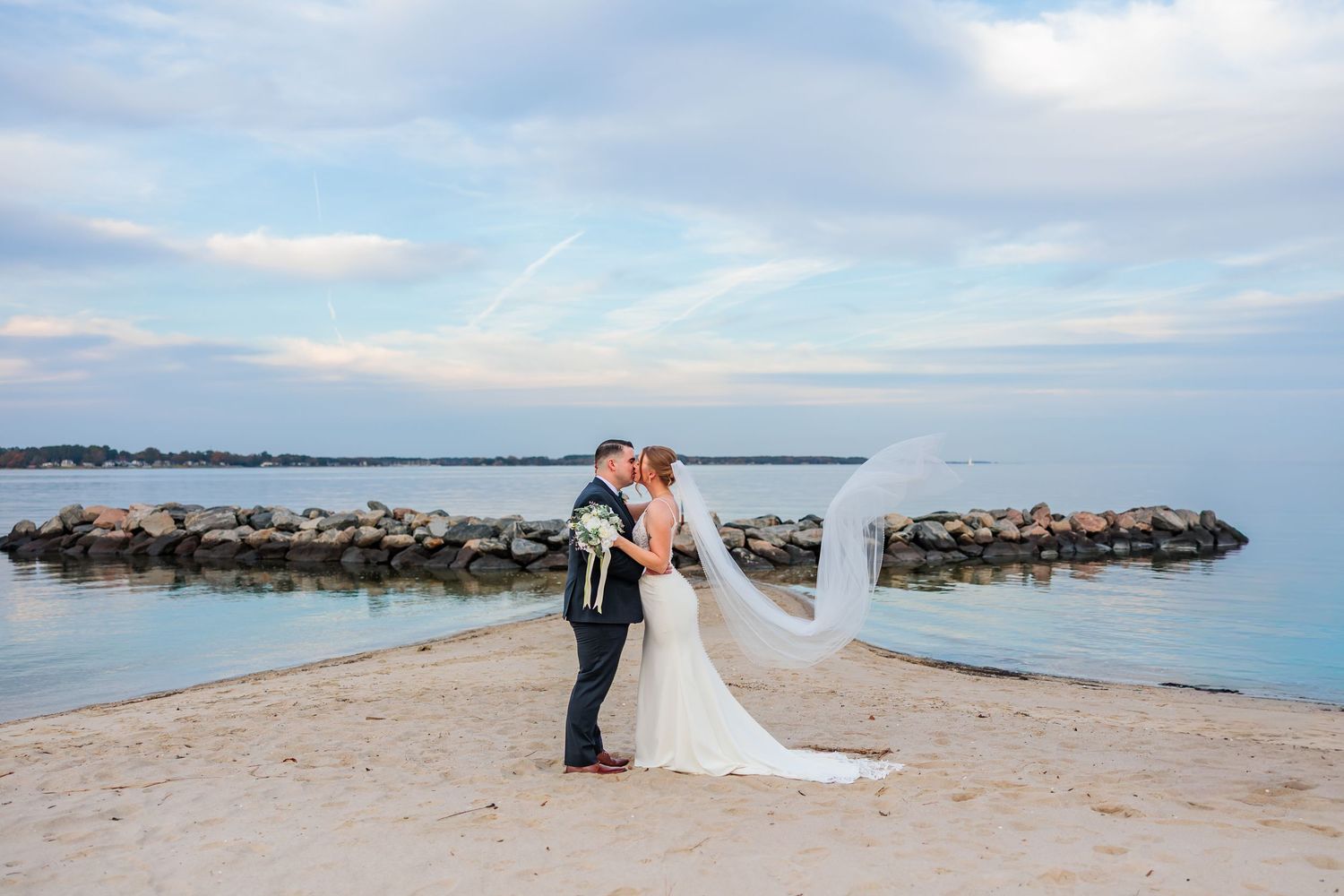 Bride and groom share a romantic kiss on a sandy beach by the ocean, with the veil flowing in the wind.