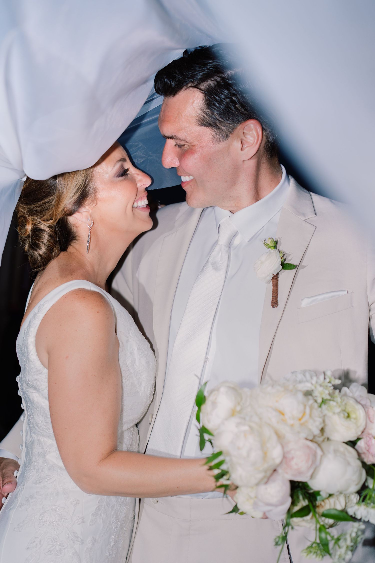 Bride and groom smiling at each other during wedding ceremony, bride holding white peony bouquet, groom in beige suit.