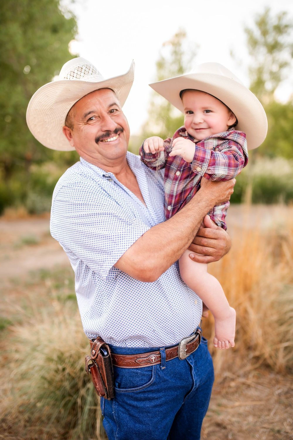 Smiling grandfather in cowboy hat holding happy baby also wearing a cowboy hat outdoors.