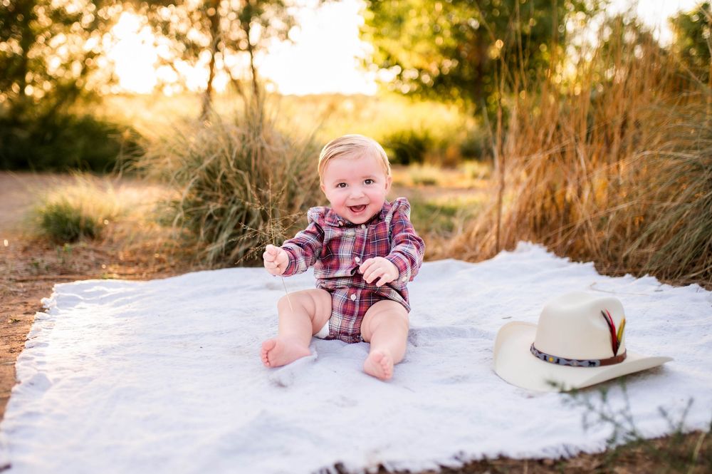 Adorable baby in plaid outfit sitting on white blanket outdoors next to a cowboy hat.