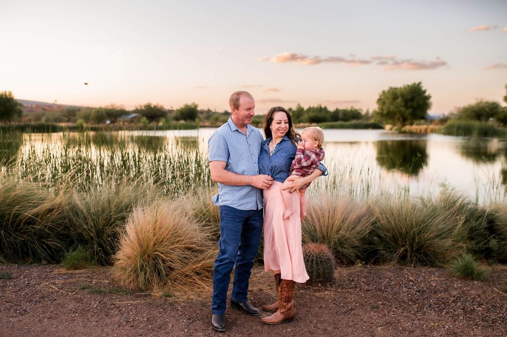 Family of three poses by a serene lake at golden hour sunset in a lush outdoor setting.