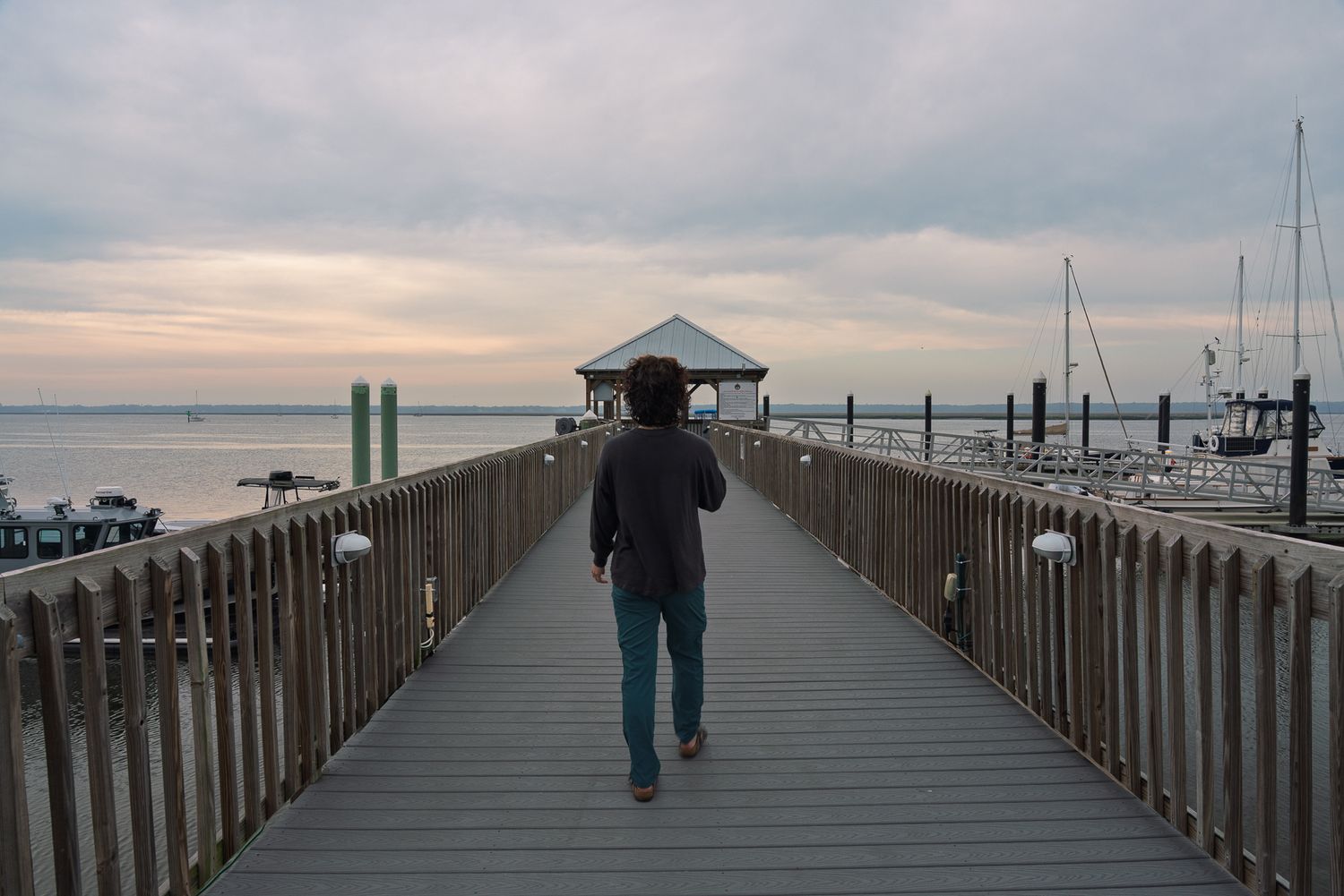 A lone person walks along a wooden pier toward a gazebo overlooking a calm harbor at dusk.
