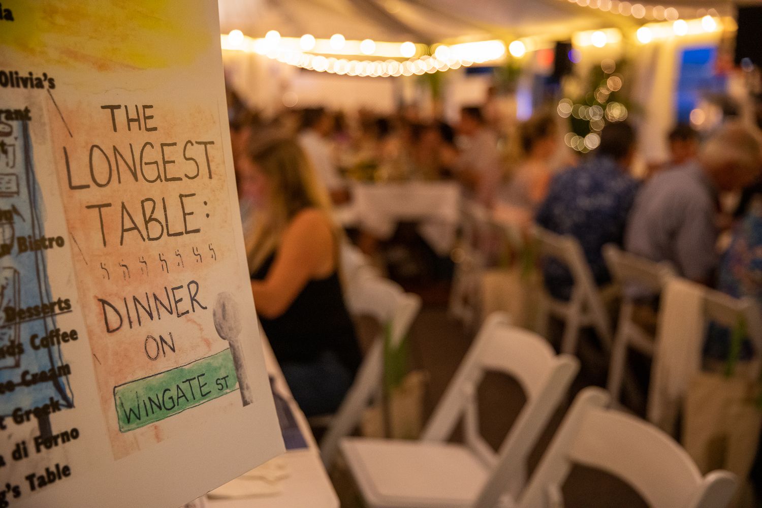 Sign for The Longest Table Dinner on Wingate St event with guests dining under string lights in background.