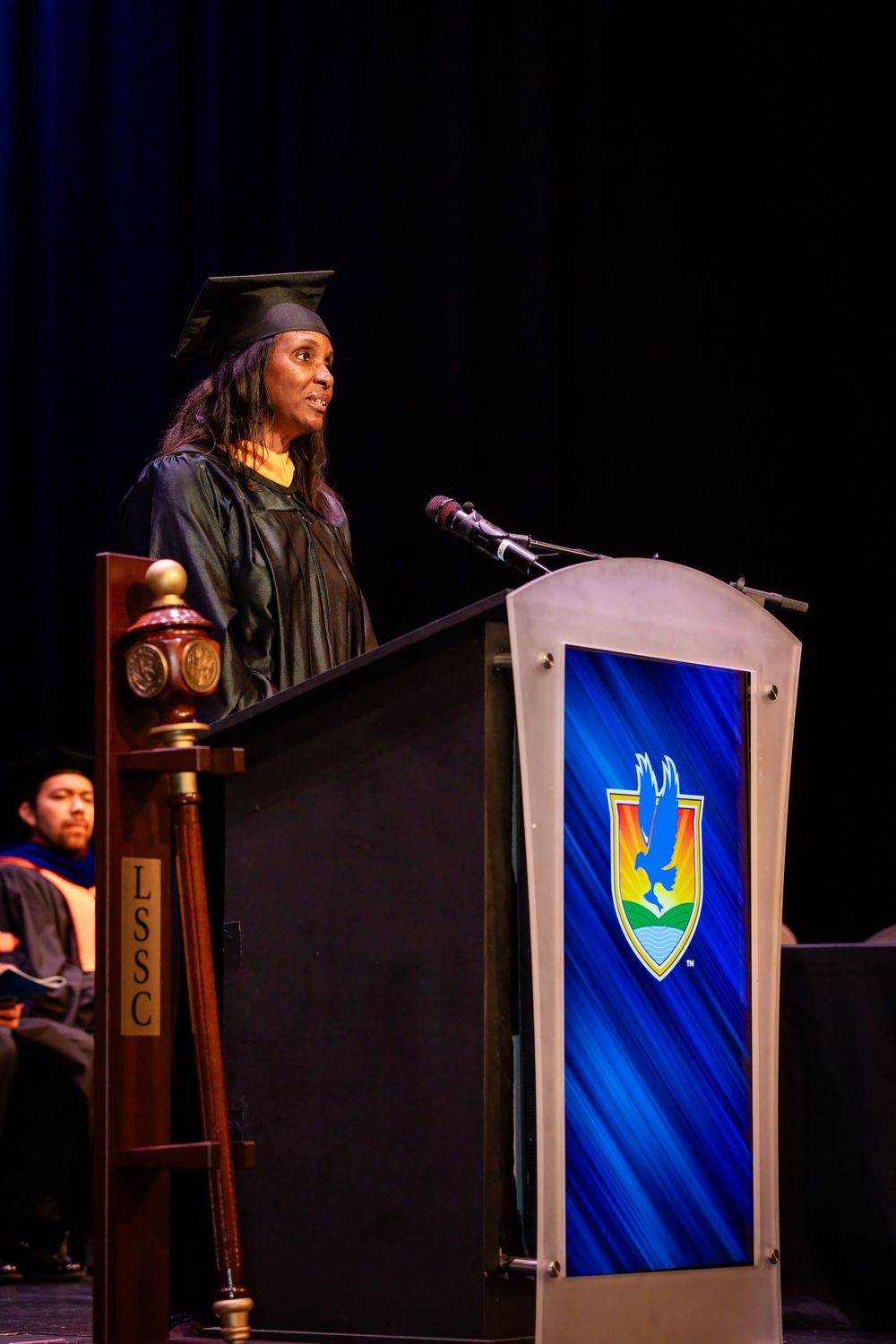 Graduate in cap and gown delivers speech at podium during college commencement ceremony on stage.