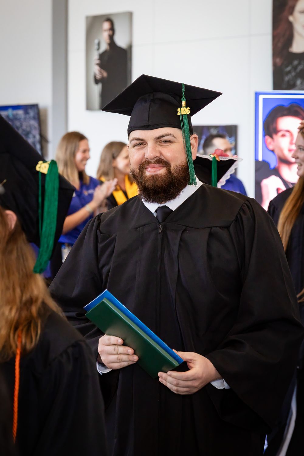 Bearded man in black graduation gown and cap holding diploma at commencement ceremony.