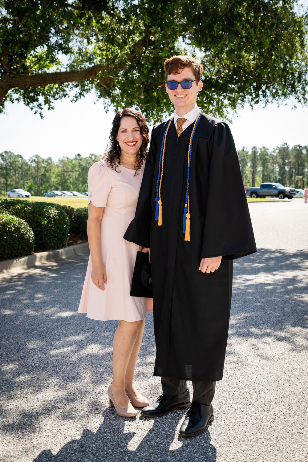 A proud graduate in black cap and gown with blue and gold tassels poses with a smiling woman in a pink dress outdoors.
