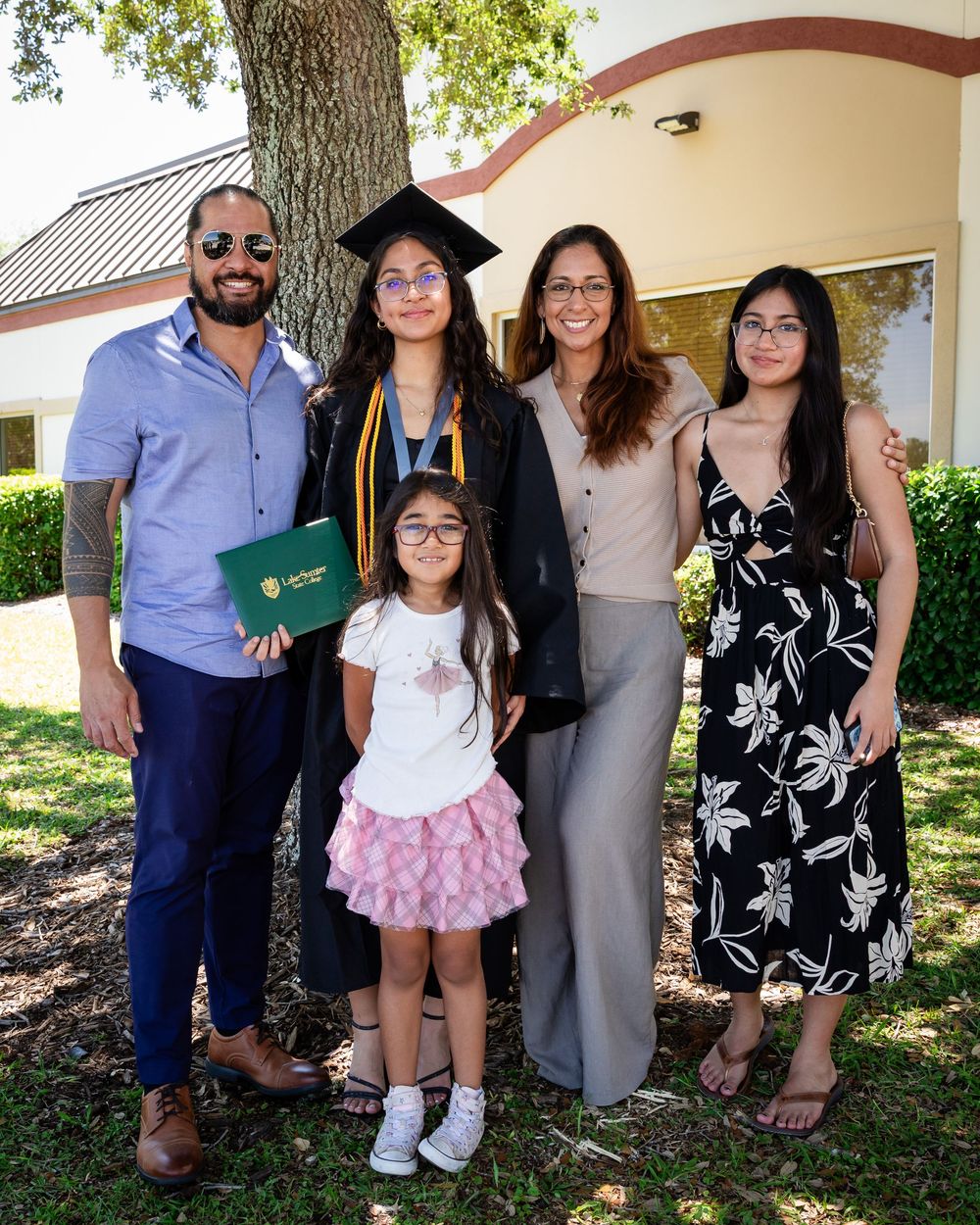 Family celebrating a graduation outdoors, graduate in cap and gown holding diploma, posing with four relatives near a tree.