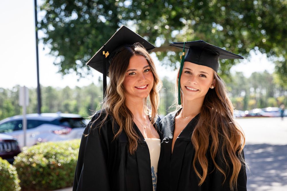 Two smiling women in black graduation caps and gowns posing outdoors on a sunny day.