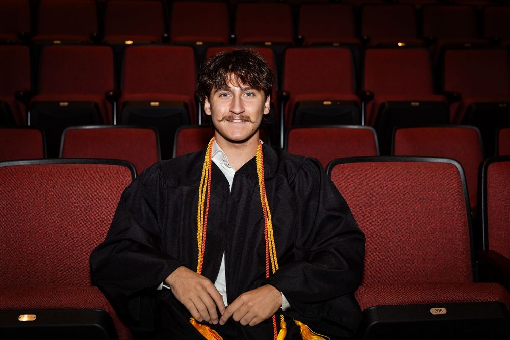 Young man in graduation gown and gold stole sitting in red auditorium seats, smiling for graduation portrait photo.