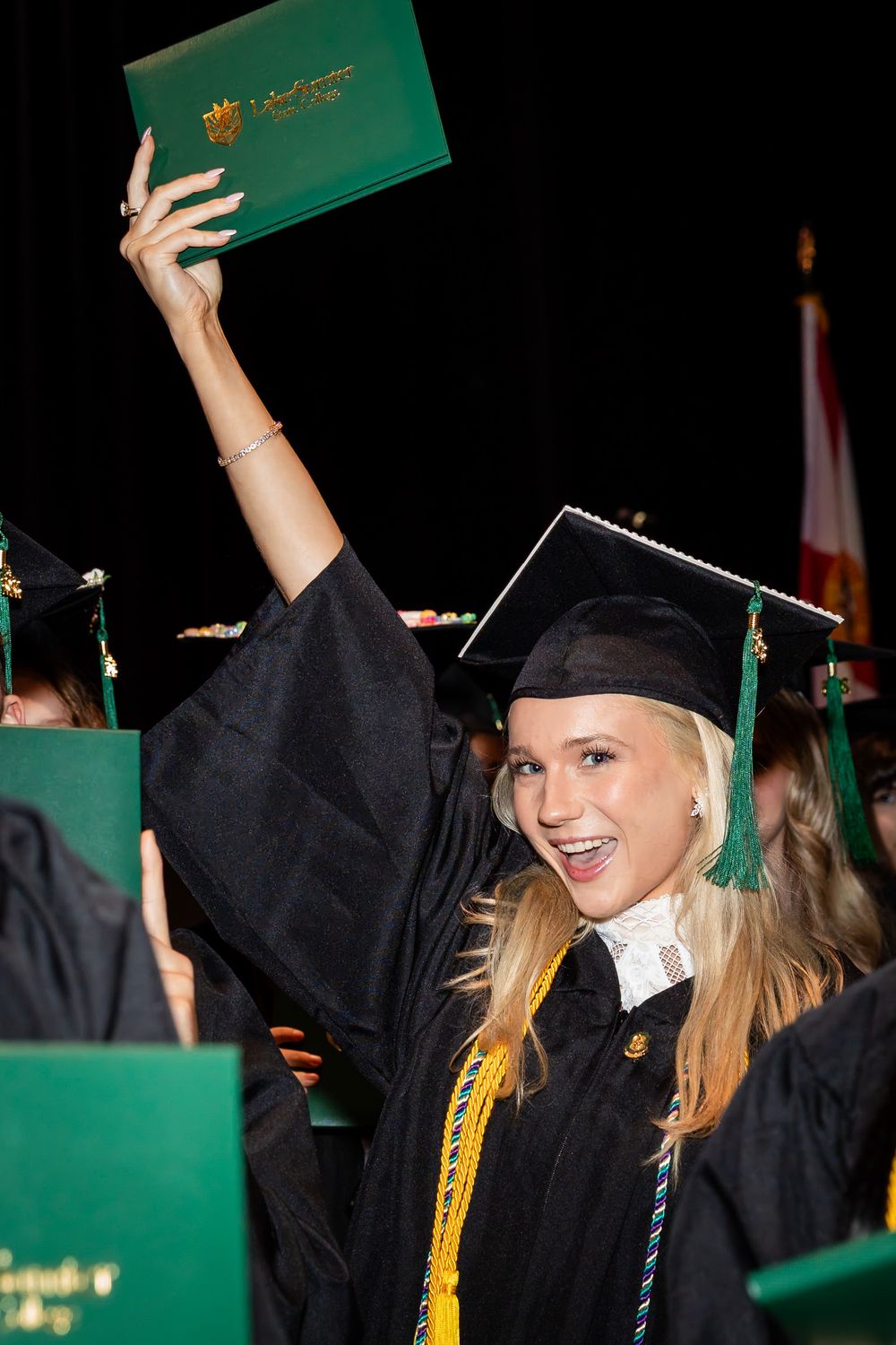 Excited female graduate in black cap and gown raises green diploma cover at university commencement ceremony.